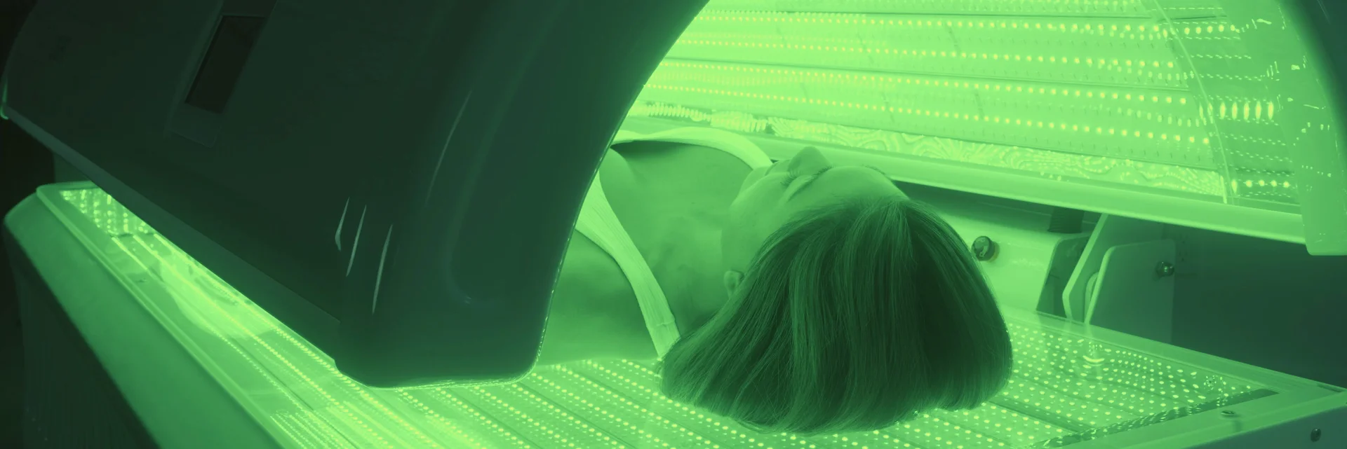 Person lying face down inside a tanning bed illuminated with green light.